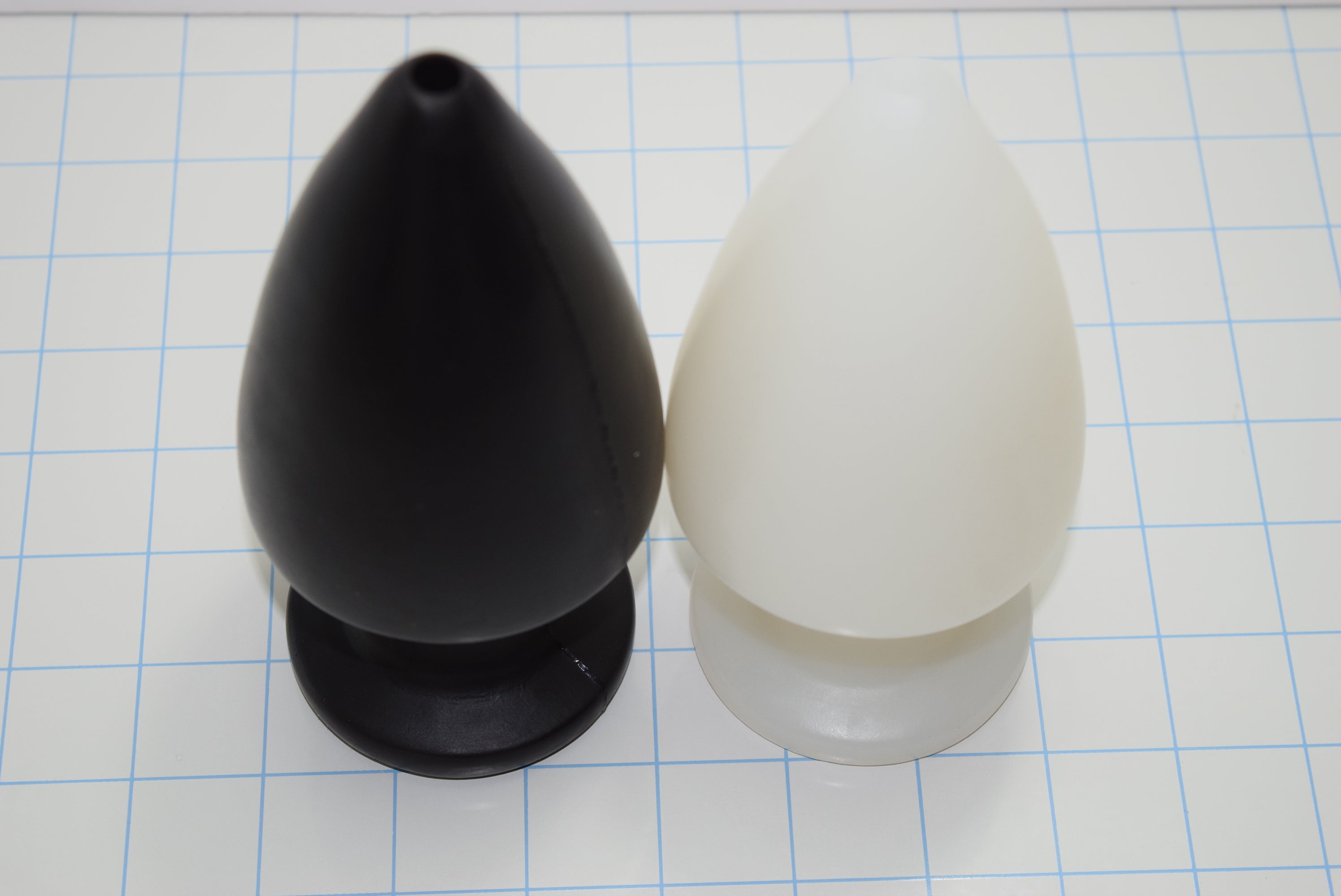 Black and white silicone retention enema nozzles standing side-by-side vertically on a blue grid background.