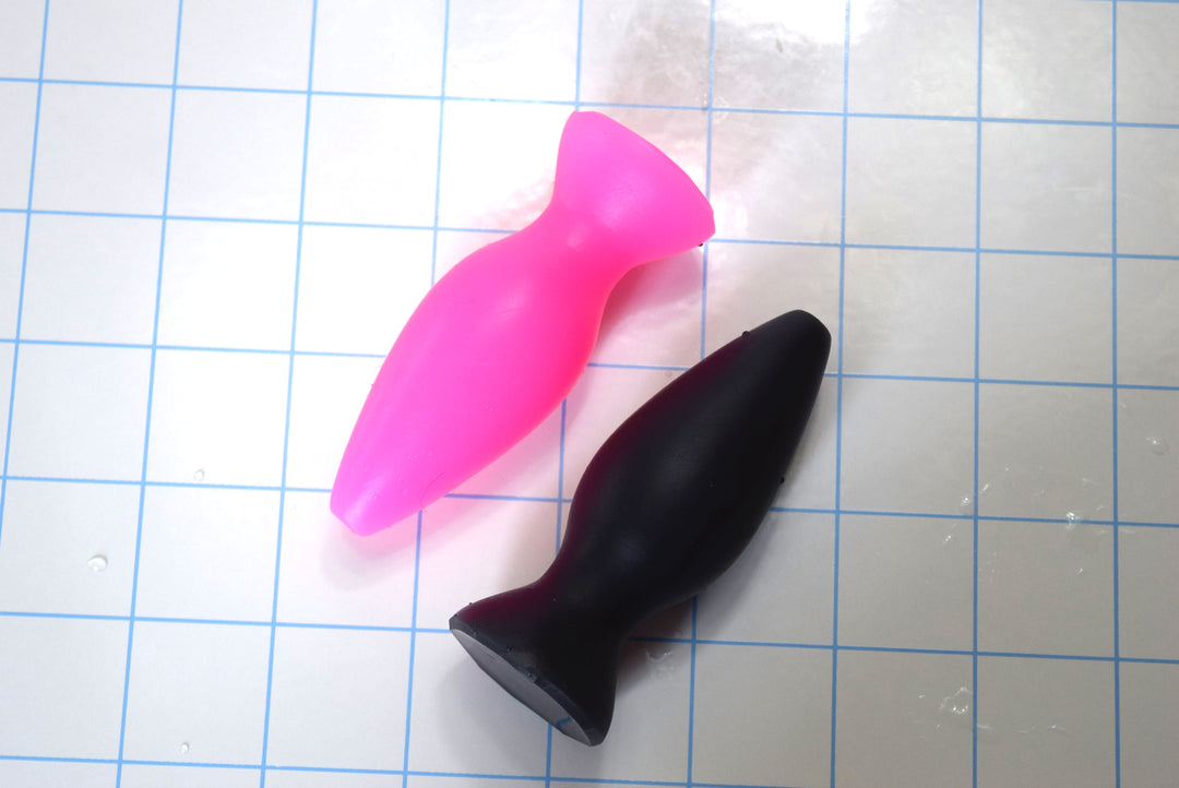 Pink and black small silicone retention plugs lying horizontally side-by-side on a blue grid background.