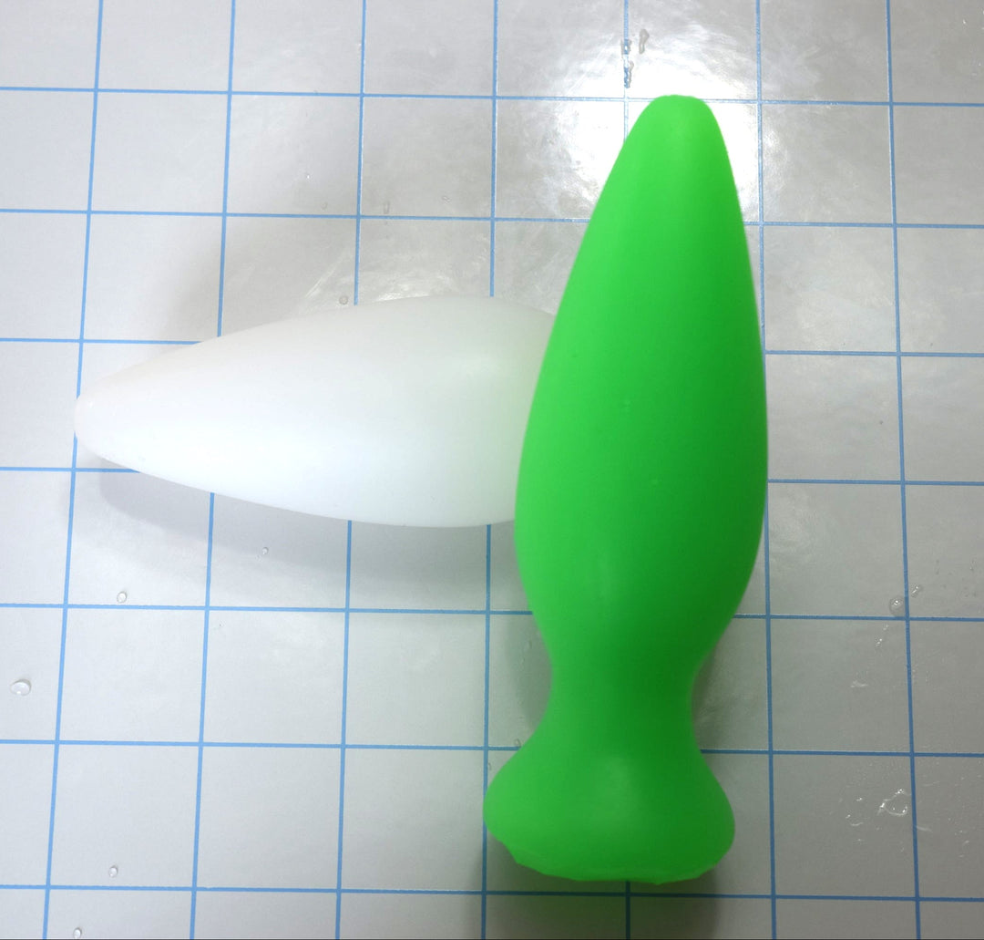 Green silicone retention enema nozzle standing upright next to a white nozzle lying on a blue grid background.
