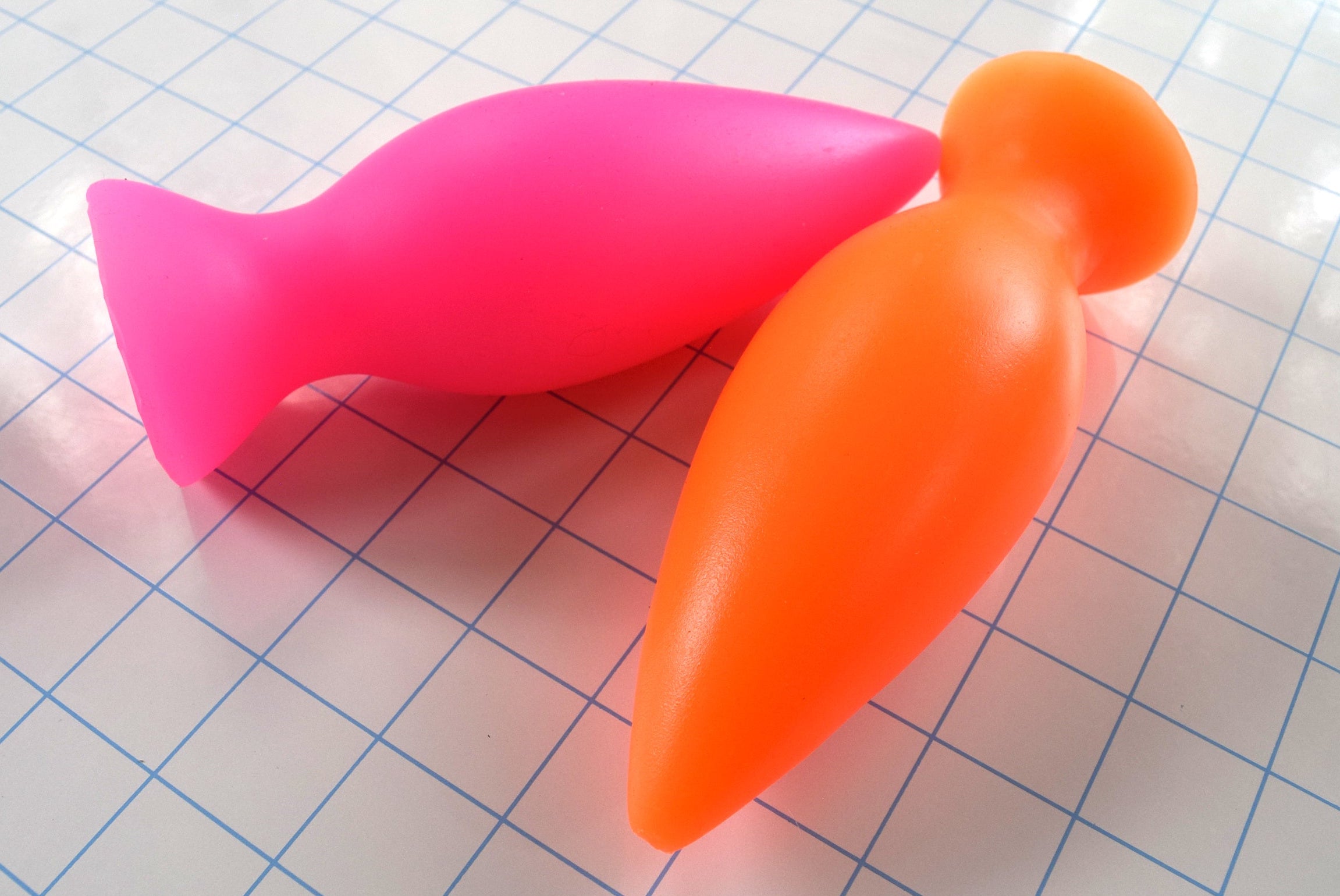 Pink and orange stubby silicone retention plugs resting side-by-side on a blue grid background.