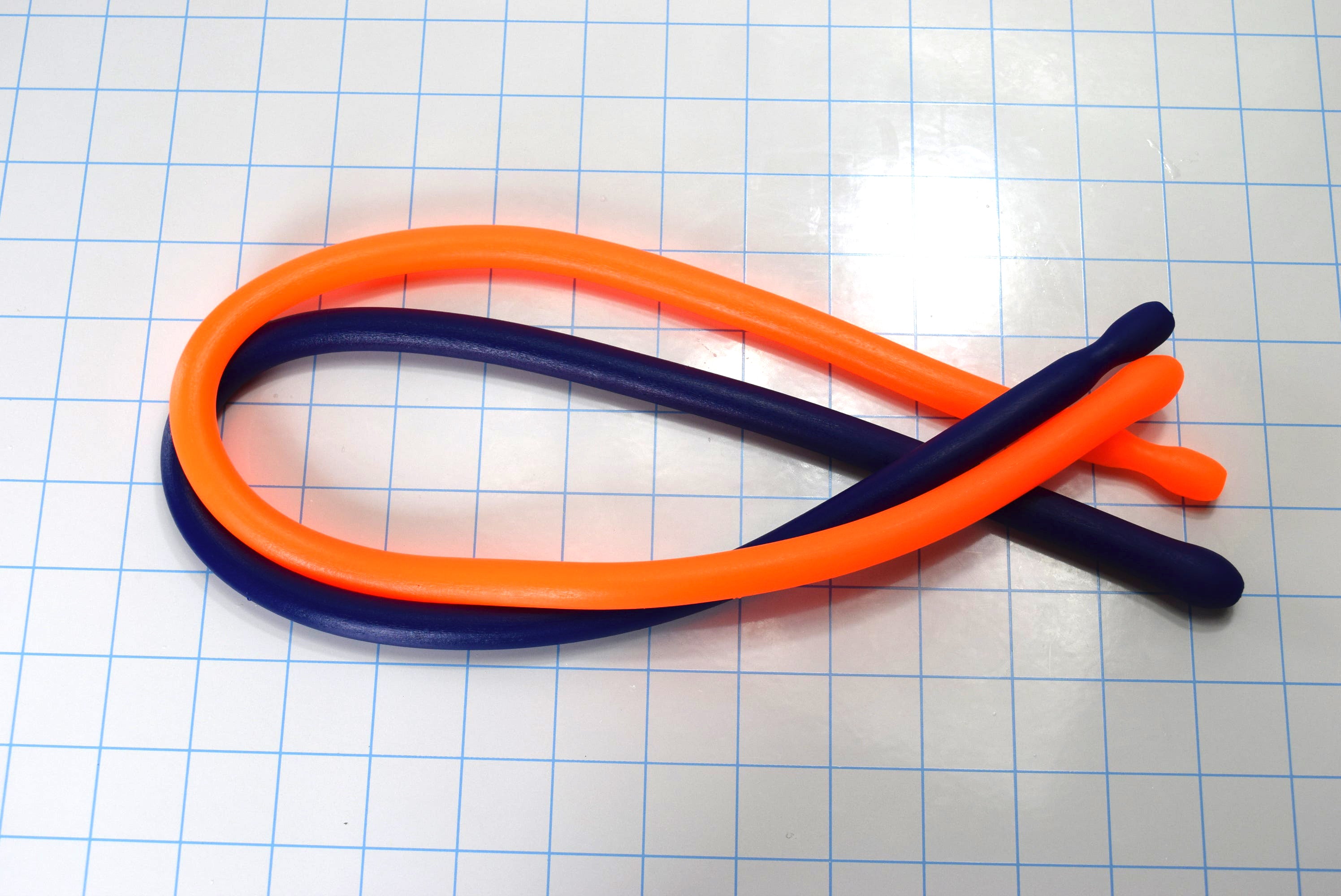 Orange and blue flexible SSND_24_BT silicone sounds looped in an oval shape on a blue grid background, featuring a 14mm egg-shaped tip, 12mm shaft, and 24-inch length.