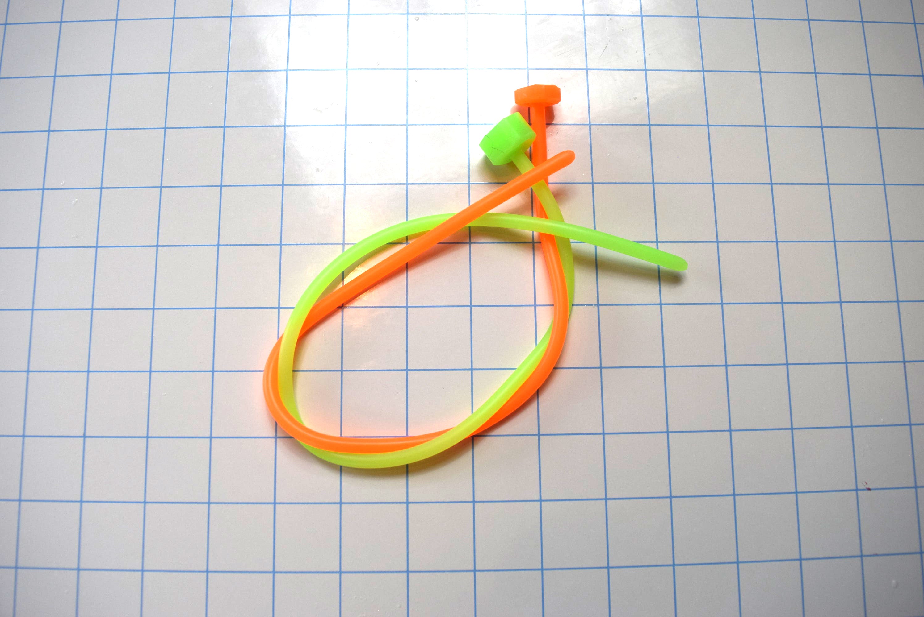 Pair of SSND-5.5mm silicone urethral sounds in fluorescent green and fluorescent orange featuring 5.5mm diameters (16 French), smooth rounded tips, solid core constructions, and 15-inch insertable lengths on a grid background.