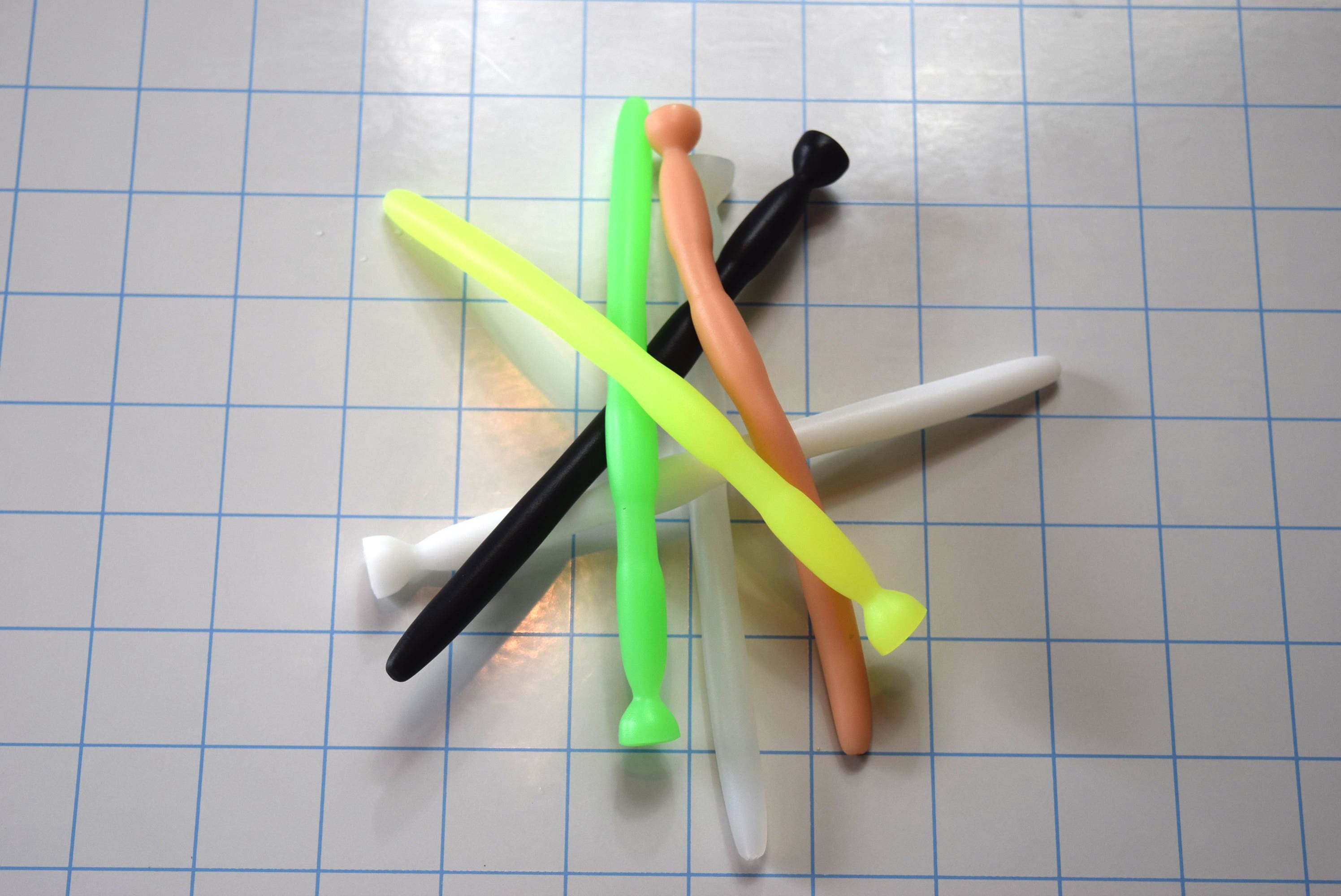 Assortment of SSND-BMK2 silicone urethral sounds in various colors (neon green, peach, black, white, translucent yellow) crossed on a blue grid background, showcasing the 8mm entry shaft, dual 11mm expansion barrels, 5.5mm retention groove, and safety flange base.
