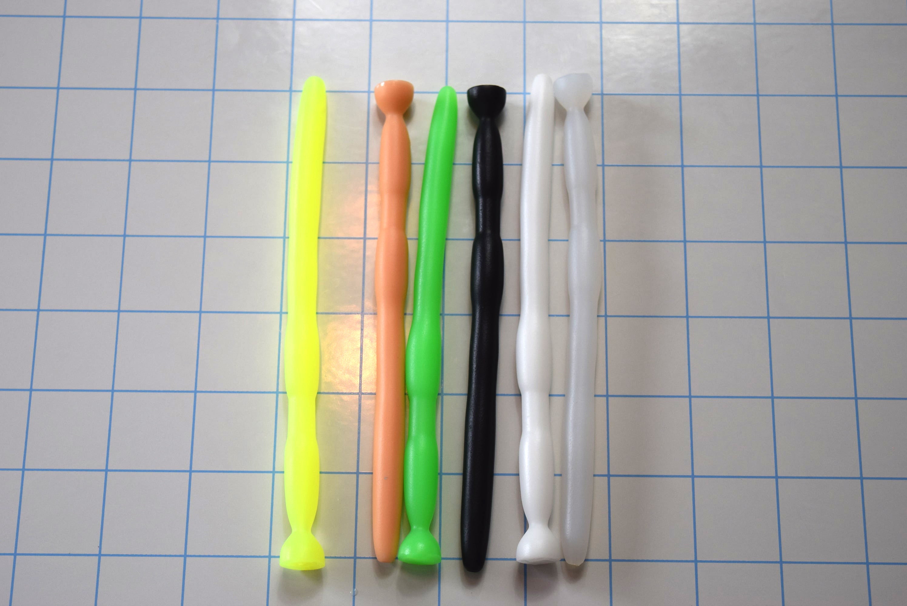 Assortment of SSND-BMK2 silicone urethral sounds in various colors (neon green, peach, black, white, translucent yellow) crossed on a blue grid background, showcasing the 8mm entry shaft, dual 11mm expansion barrels, 5.5mm retention groove, and safety flange base.