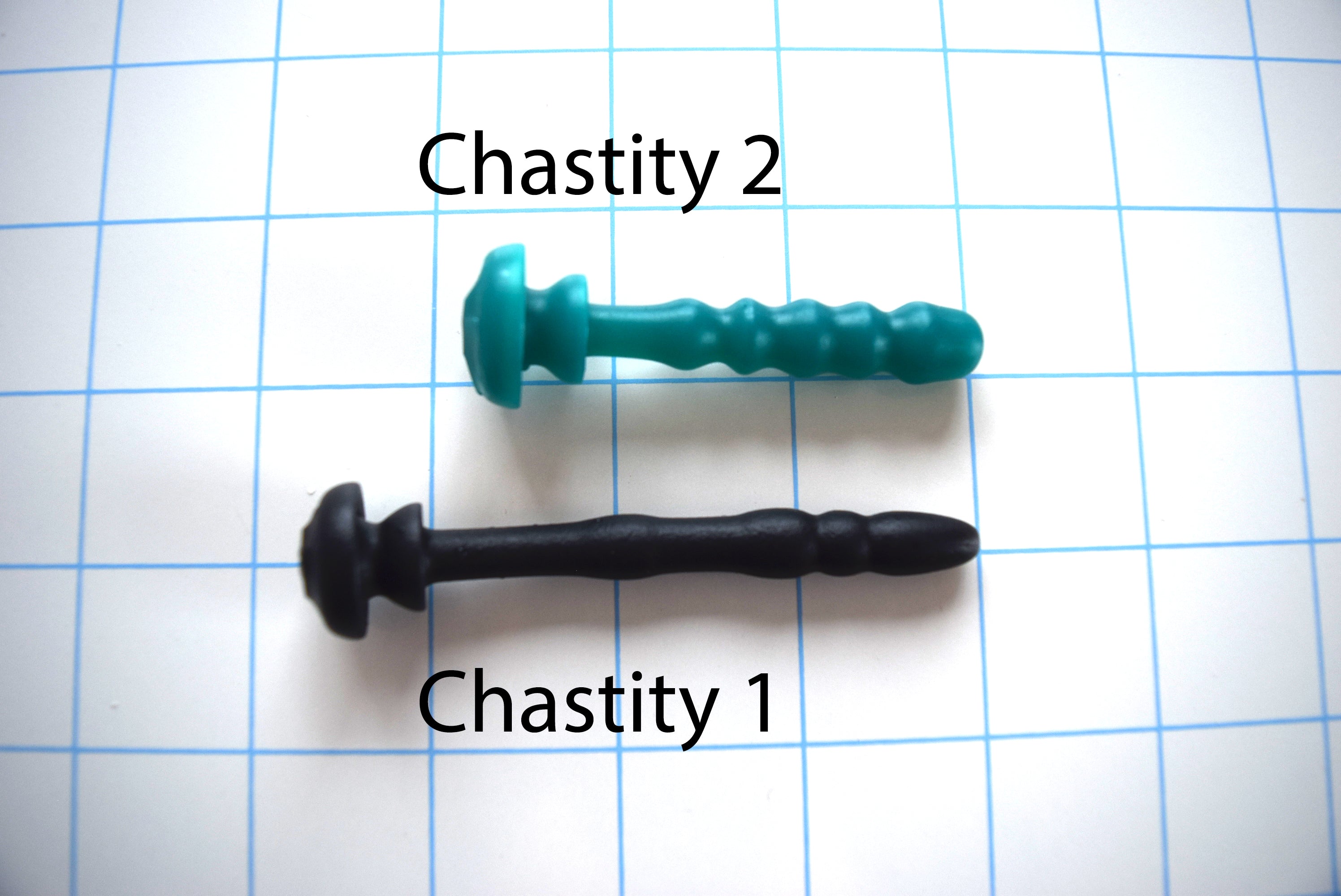 Side-by-side comparison of the green Chastity 2 silicone urethral plug insert and the longer black Chastity 1 insert on a grid background.