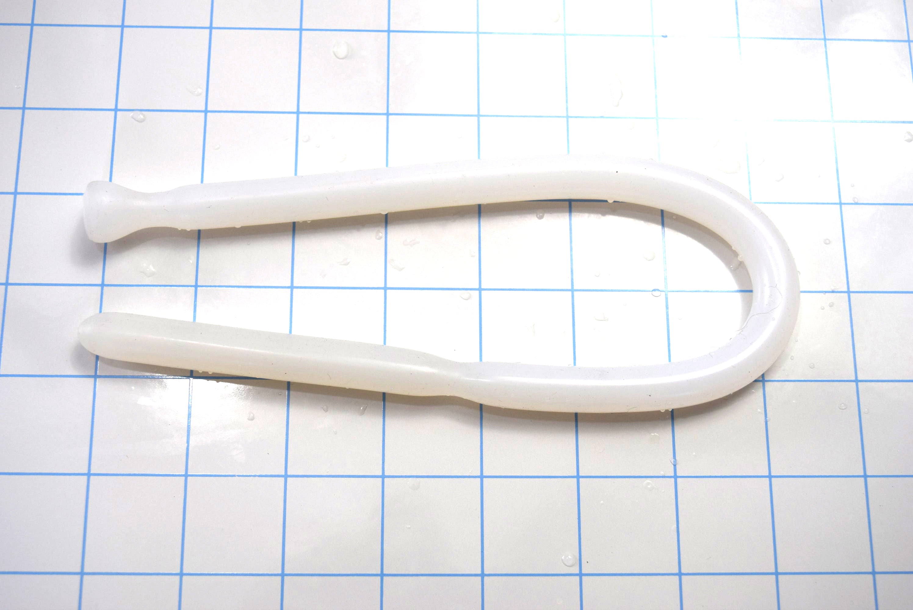 Translucent white SSND-DS 9-12 flexible silicone sound bent in a U-shape on a blue grid mat, displaying its grooved tip and flanged safety base.