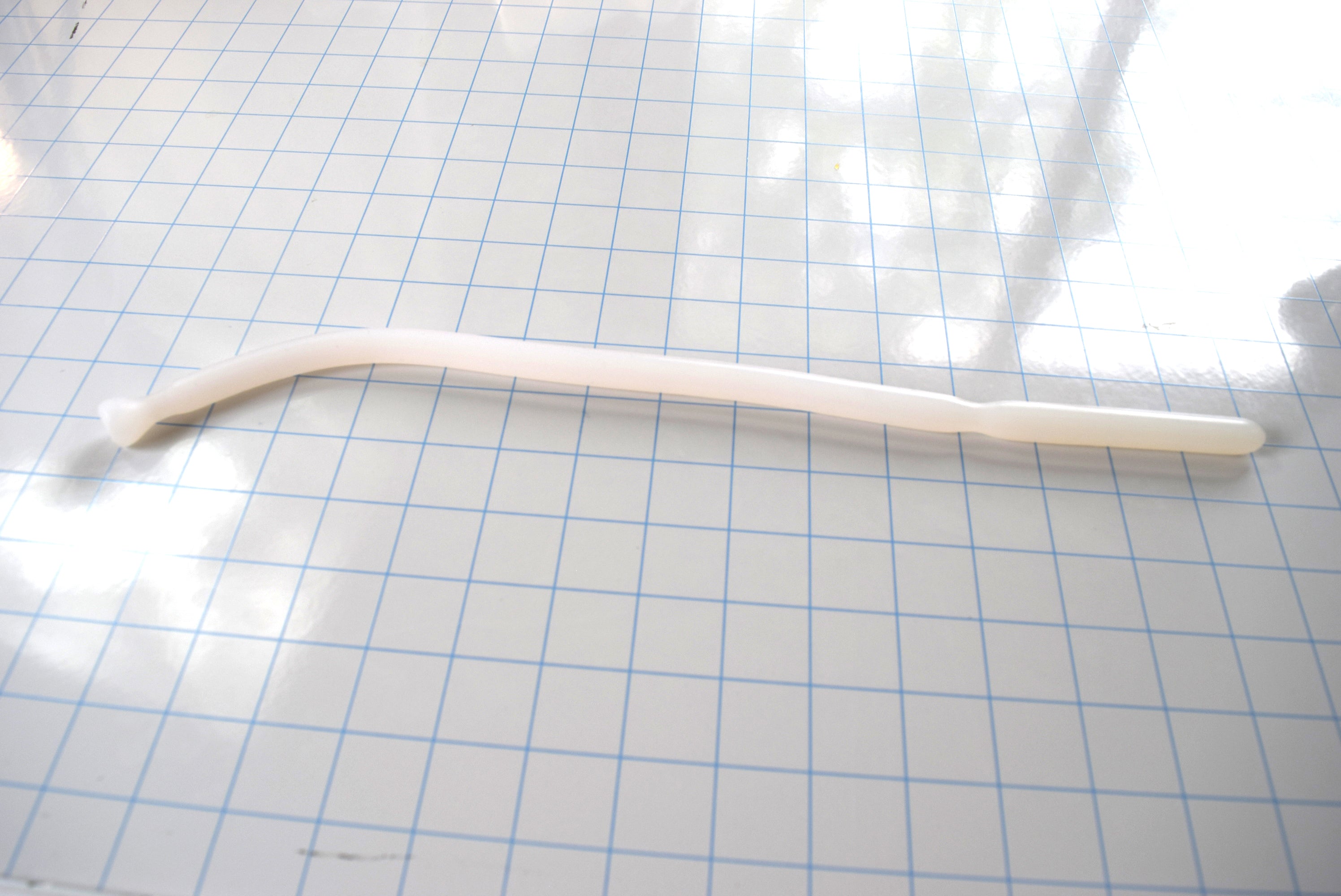 Translucent white SSND-DS 9-12 silicone sound featuring a grooved tip, 12mm shaft, and flanged safety base laid horizontally on a blue grid background.