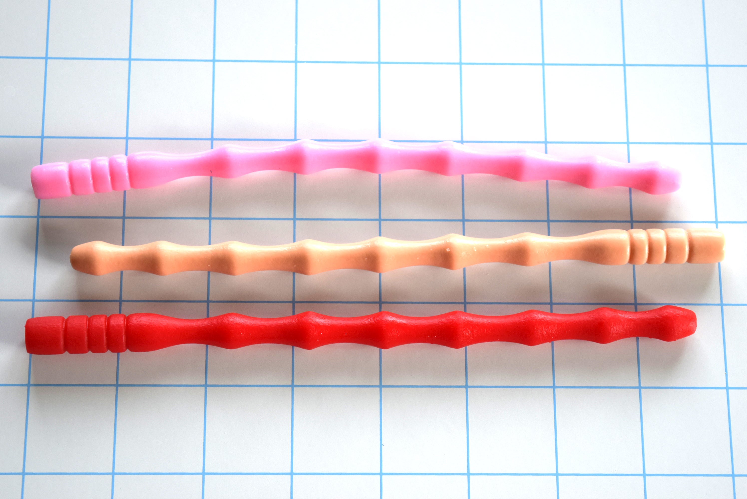 SSND-GARTER urethral sounds displayed horizontally in pink, peach, and red variants on a grid background highlighting the 7mm tapered tip, 6 contoured wave segments peaking at 10.5mm, and the distinctive 3-barrel rib safety base flange.