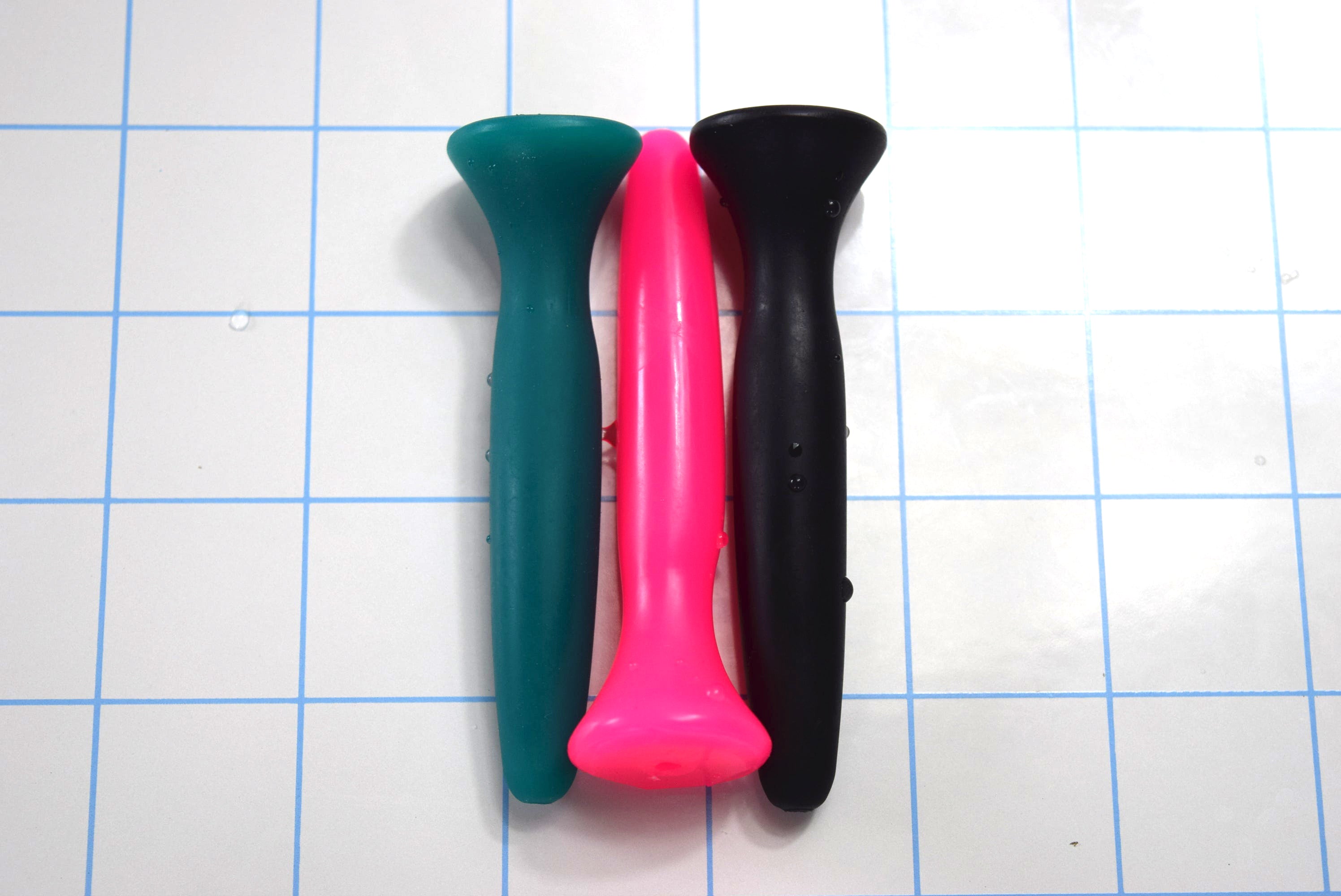 SSND-JWW 6 silicone urethral sound shown vertically in three color choices—teal green, bright pink, and black—on a white surface with a 1"x1" blue grid layout, displaying the 13.5mm peak diameter shaft, 11mm retention valley, and flared safety flange base.