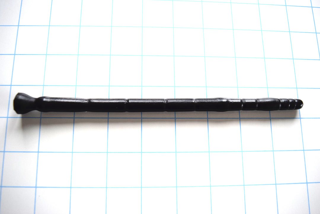 SSND-MLTIDML 8-12 intermediate black silicone urethral sound shown horizontally on a white background with 1"x1" blue grid squares from Screenshot 2026-07-02 160647.jpg, highlighting its graduated micro-segments near the 7mm tip, 12mm peak stretch barrel, and 8-inch insertable length.