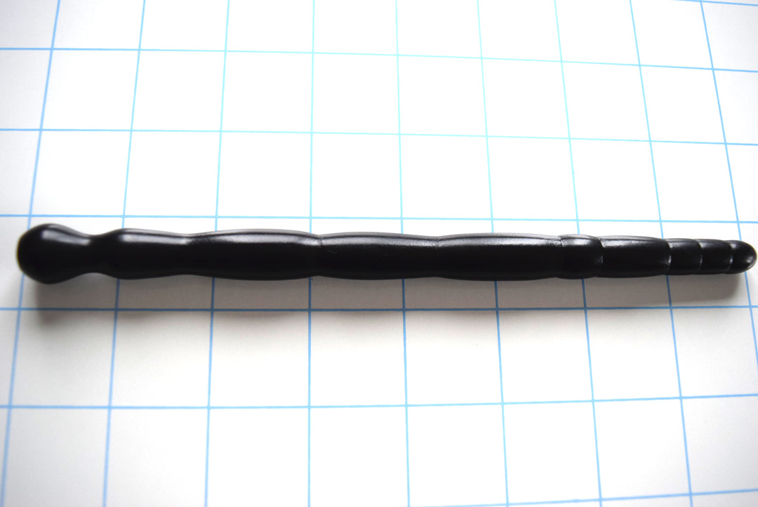 SSND-MLTIDML 9-13mm multi-dimensional black silicone urethral sound resting on a grid background, highlighting its 9.5mm rounded tip, progressive barrel-style segments, 9.3mm retaining valley, and unique ball-shaped safety flange.