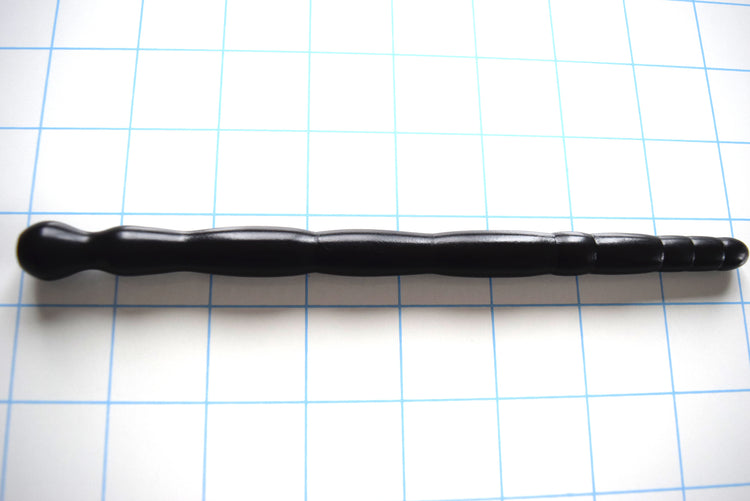 SSND-MLTIDML 9-13mm multi-dimensional black silicone urethral sound resting on a grid background, highlighting its 9.5mm rounded tip, progressive barrel-style segments, 9.3mm retaining valley, and unique ball-shaped safety flange.