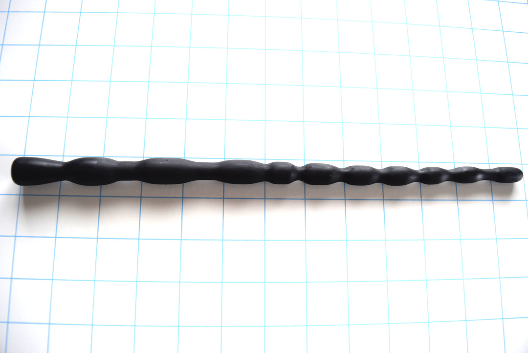 SSND-MRB 7-16 graduated silicone sounding rod in matte black shown laid out horizontally on a white background with 1-inch blue grid lines, featuring 10 expanding ribs up to 16.4mm and a flared safety base flange.
