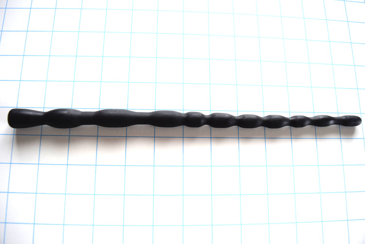 SSND-MRB 7-16 graduated silicone sounding rod in matte black shown laid out horizontally on a white background with 1-inch blue grid lines, featuring 10 expanding ribs up to 16.4mm and a flared safety base flange.