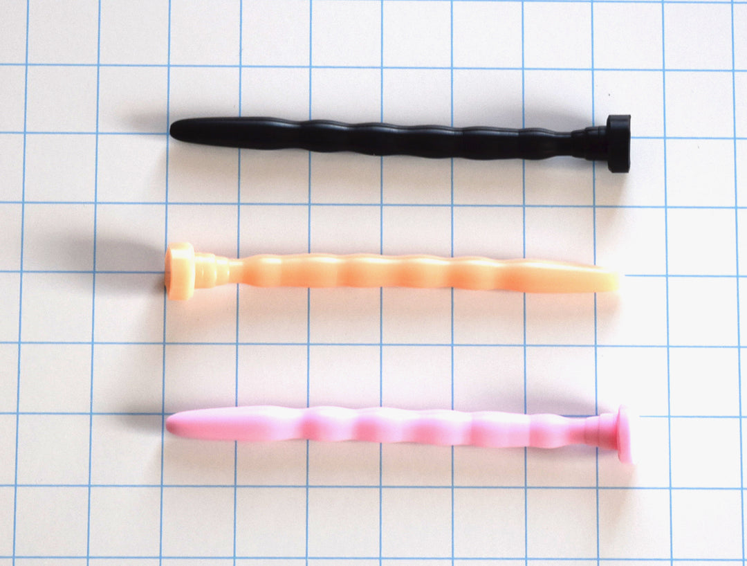 SSND-RIPPLE 8-12 wave-contoured silicone urethral sound shown in black, flesh, and pink variants on a grid background, highlighting the five undulating ripple crests peaking at 12mm, 5.5-inch insertable length, retention valley, and safety base flange