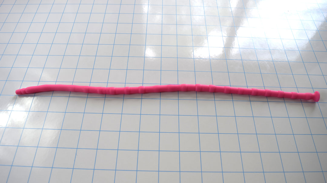 Bright pink SSND-RNDMBALL 7-10mm silicone sound with a multi-ball ribbed shaft and flanged base lying horizontally across a blue grid background.