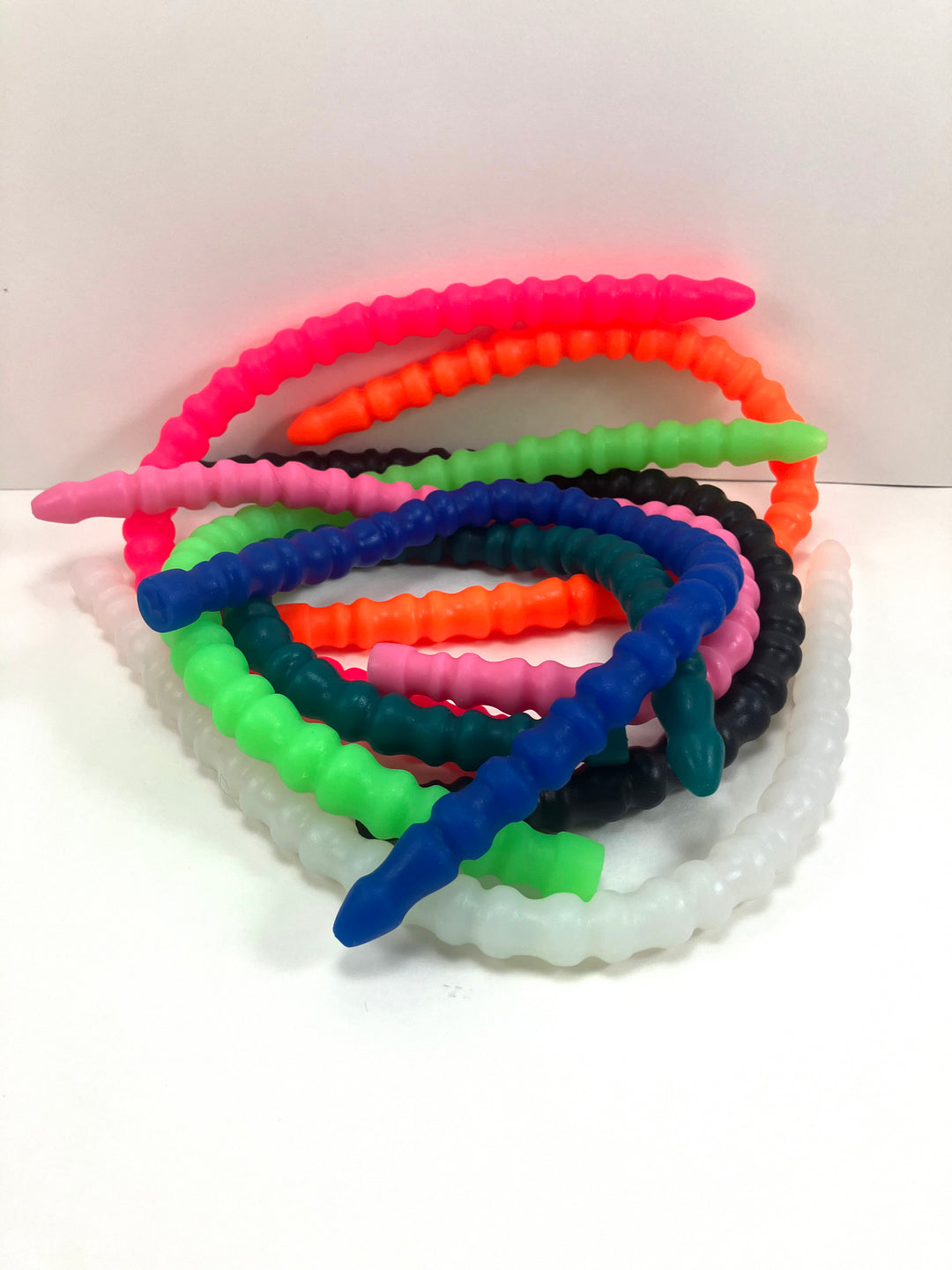 A collection of SSND-RNDMRIB silicone urethral sounds in various colors including pink, orange, green, blue, black, and translucent white, featuring alternating ribbed sections and safety flange bases on a white background.
