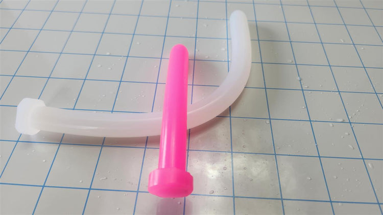 Bright pink 14mm SSND silicone sound standing vertically next to a curved translucent white silicone rod on a blue grid surface.