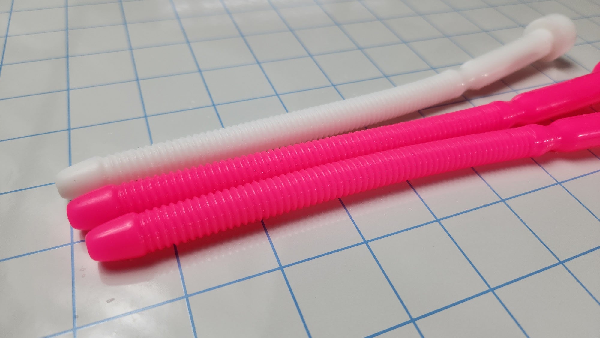 Two hot pink and one white SSND-THREADED13 silicone urethral sounds resting on a blue grid background, featuring an 8.3-inch insertable length, threaded shaft, and retention valley.