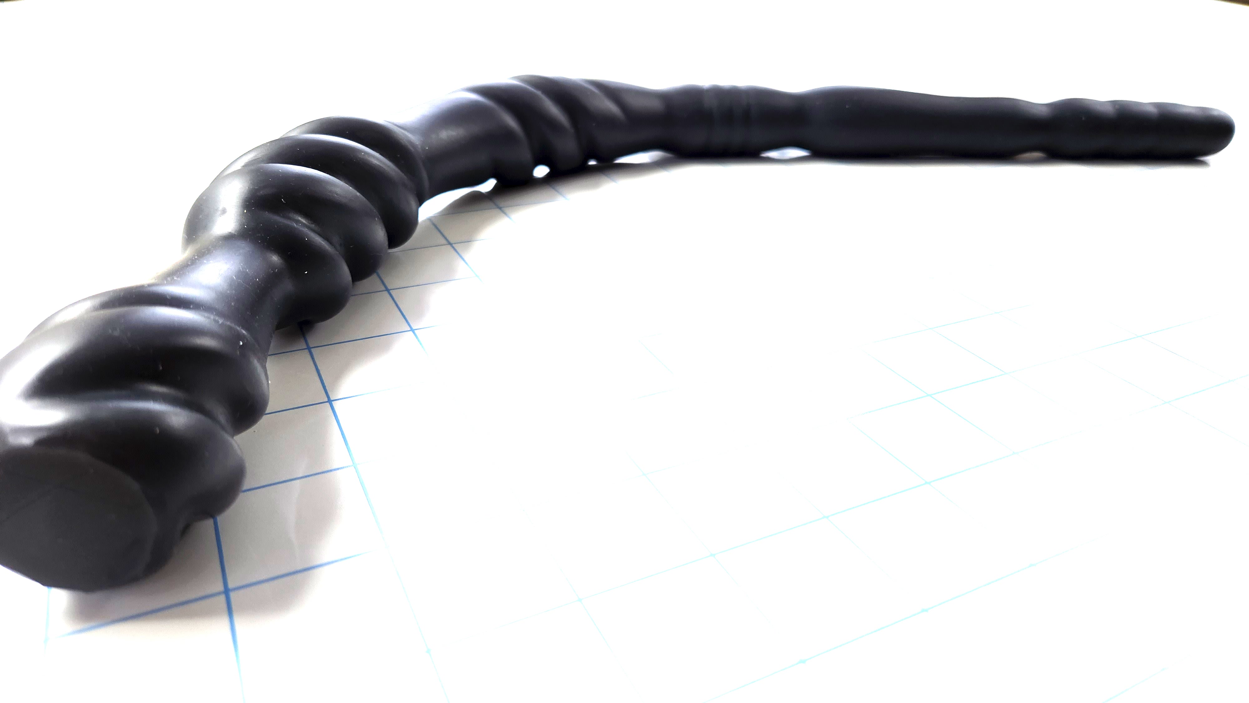 Black Triple Spiral 22-inch silicone nozzle featuring intricate spiraled textures and a graduated design for varied stimulation on a white grid background, showing the flange in focus