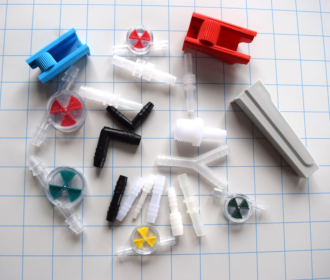 Assortment of tubing accessories including roller clamps, flow wheels, connectors, adapters, and elbows on a blue grid background.